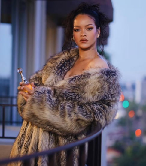 Rihanna posa em varanda usando casaco de pele e segurando frasco de perfume.