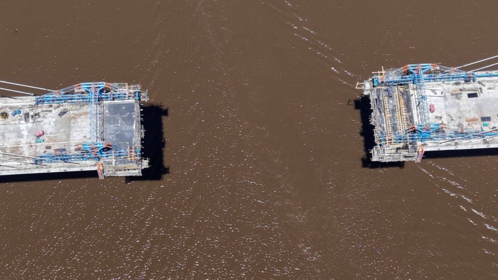 Vista aérea mostra dois lados da ponte em construção avançando sobre o rio. Vista aérea mostra dois lados da ponte em construção avançando sobre o rio.