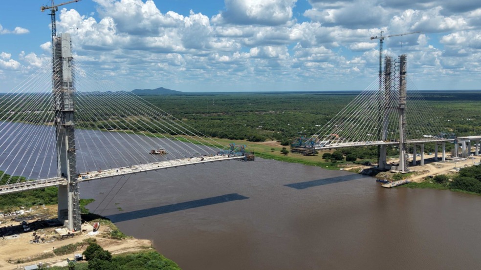 Ponte estaiada em construção atravessa grande rio cercado por vegetação.