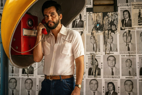 Homem usa telefone público diante de parede coberta por cartazes políticos antigos.